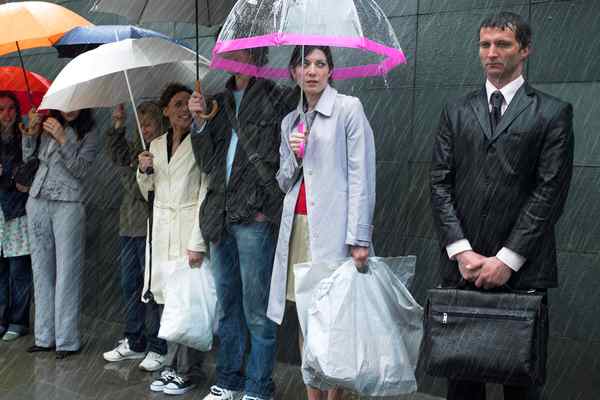 A group of people stand outside in the rain. A few of them have umbrellas.