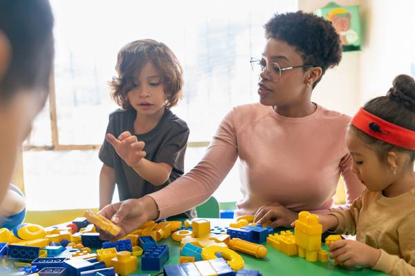 A preschool teacher and several children play with toy blocks in a classroom.