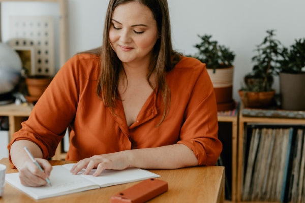 A woman sitting at a table writing a to-do list in a notebook.