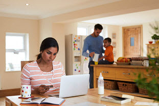 A woman works on a laptop while a man and a child are shown in the background. 