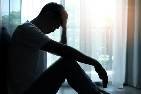 A depressed man sits on the floor while holding his head in his hand.