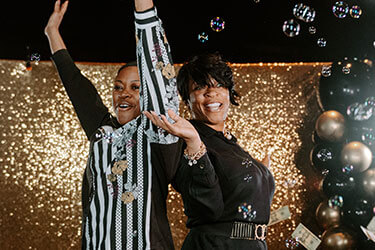 Two smiling Purdue Global graduates dancing in front of a gold background, surrounded by black and gold ballooons and bubbles